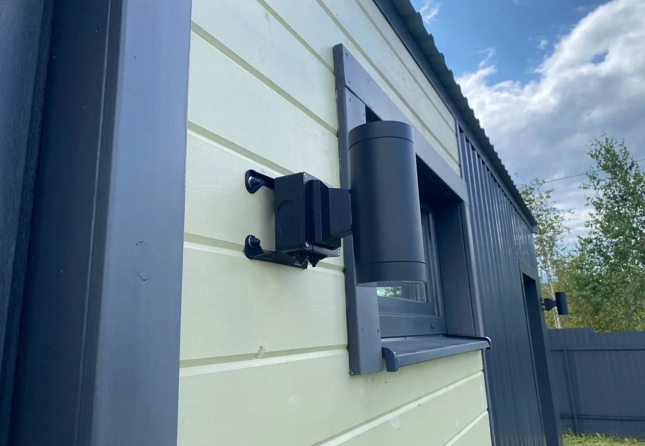 Коробка BOXFORCAM™ на стене из светлого сайдинга рядом с цилиндрическим настенным светильником